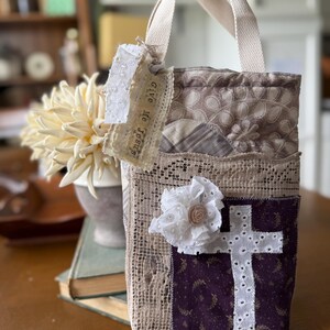 Può includere: Una borsa tote fatta a mano con manico color crema e design patchwork. La borsa presenta una croce bianca su sfondo viola, un fiore bianco e un'etichetta con la scritta "Give Me Jesus".