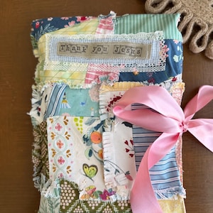 May include: A patchwork fabric journal with a pink ribbon closure. The journal is made of various patterned fabrics in shades of blue, green, yellow, and floral prints. The text "Thank you Jesus" is displayed on a small fabric label.