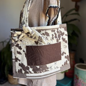 May include: A handmade tote bag with a cowhide pattern, brown accents, and a decorative pocket. The bag features a cream-colored floral patterned strap and a cream-colored lace detail. The bag is designed with a patchwork style.
