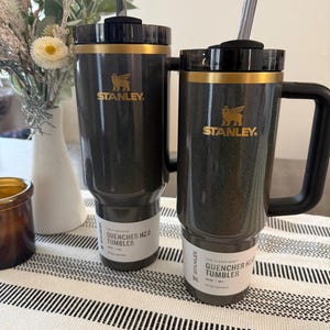 Może przedstawiać: Dwa czarne kubki Stanley Quencher H2.0 ze złotymi akcentami i słomkami. Kubki mają złote logo Stanley i napis "QUENCHER H2.0 TUMBLER" na etykiecie. Jeden kubek ma uchwyt. Stoją na paskowanym bieżniku.