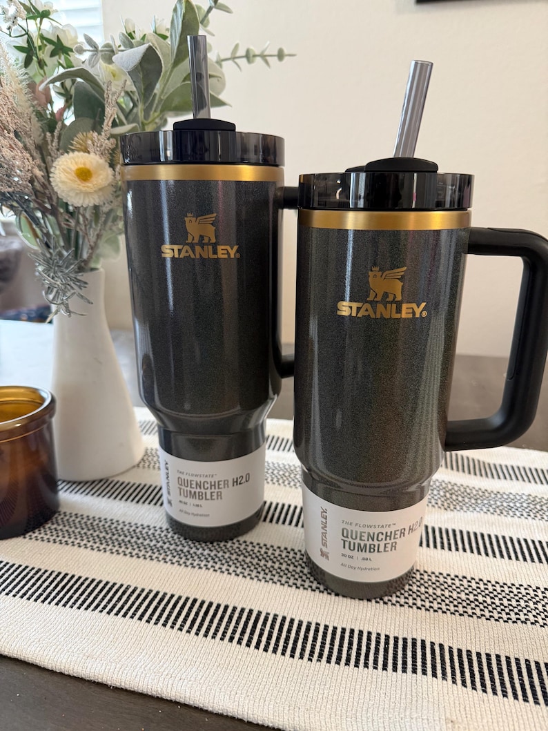 Może przedstawiać: Dwa czarne kubki Stanley Quencher H2.0 ze złotymi akcentami i słomkami. Kubki mają złote logo Stanley i napis "QUENCHER H2.0 TUMBLER" na etykiecie. Jeden kubek ma uchwyt. Stoją na paskowanym bieżniku.