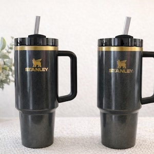 Może przedstawiać: Dwa czarne kubki Stanley ze złotymi akcentami i uchwytami. Każdy kubek ma słomkę i złote logo Stanley. Kubki stoją na białej powierzchni z wazonem kwiat&oacute;w w tle.