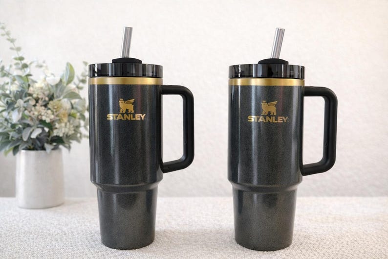 Może przedstawiać: Dwa czarne kubki Stanley ze złotymi akcentami i uchwytami. Każdy kubek ma słomkę i złote logo Stanley. Kubki stoją na białej powierzchni z wazonem kwiat&oacute;w w tle.