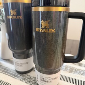 Może przedstawiać: Dwa ciemnoszare kubki Stanley Flowstate Quencher H2.0 ze złotymi akcentami i czarnymi uchwytami. Każdy kubek ma złote logo Stanley i etykietę z napisem "The Flowstate Quencher H2.0 Tumbler". Kubki znajdują się na paskowanej powierzchni.