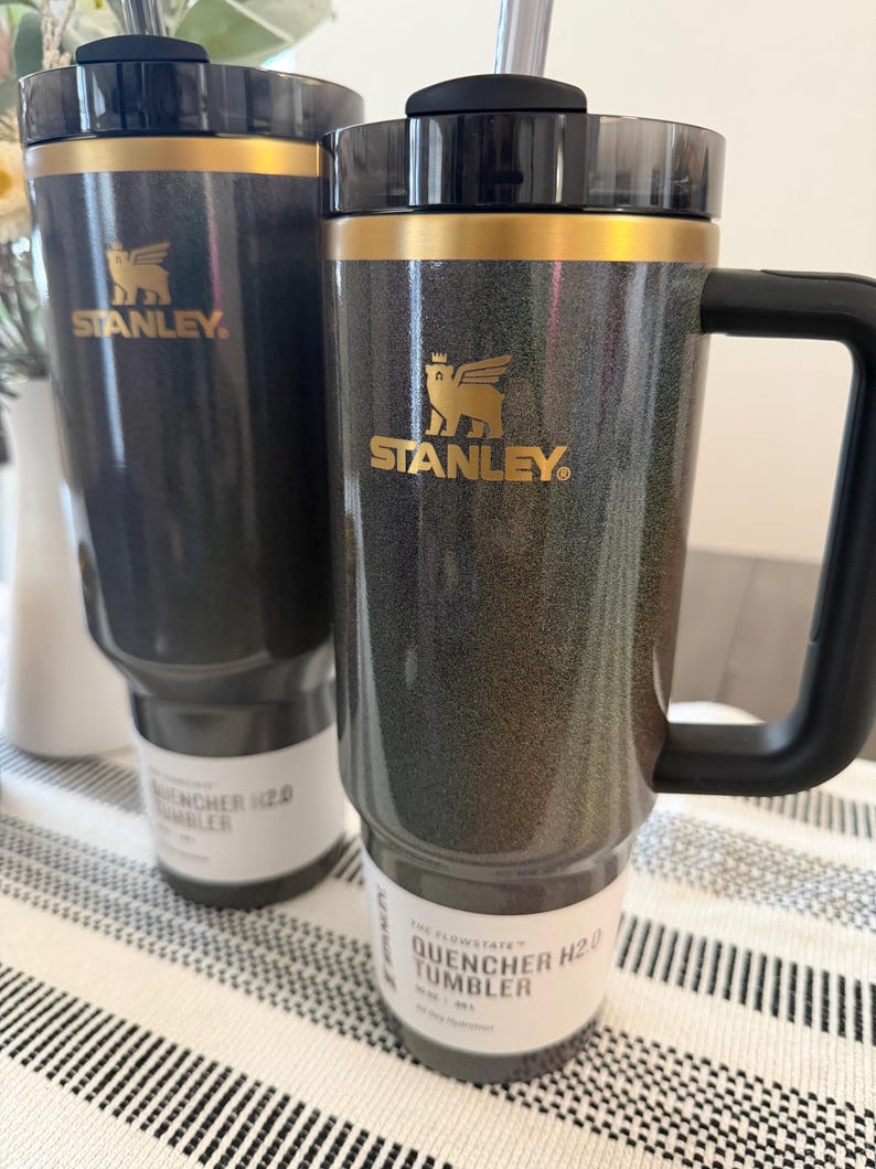 Może przedstawiać: Dwa ciemnoszare kubki Stanley Flowstate Quencher H2.0 ze złotymi akcentami i czarnymi uchwytami. Każdy kubek ma złote logo Stanley i etykietę z napisem "The Flowstate Quencher H2.0 Tumbler". Kubki znajdują się na paskowanej powierzchni.