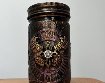 May include: A small, black glass jar with a gold lid and a gold and purple design. The jar features a gold winged cross with a rhinestone in the centre.