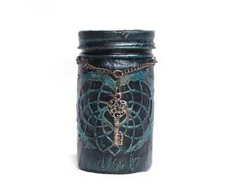 May include: A teal blue glass jar with a decorative metal key charm hanging from the lid. The jar has a distressed, vintage look and is decorated with a lace-like pattern.