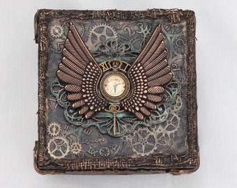 May include: A square, dark brown box with a distressed, textured surface. The box is decorated with a clock face, gears, and a pair of bronze wings.