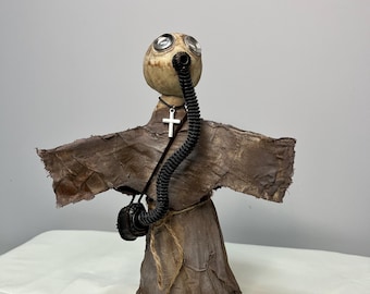 May include: A handmade doll wearing a brown robe with a gas mask and a silver cross necklace. The doll has a textured, paper mache-like appearance.