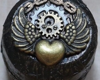 May include: A small, round, black box with a gold heart, wings, gears, and chain design. The box is decorated with a steampunk theme.