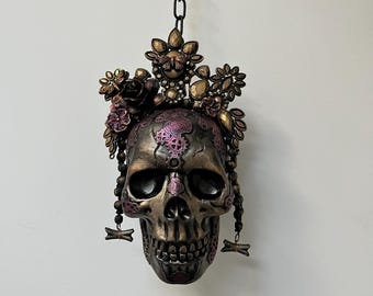 May include: A decorative skull with a floral crown and dangling beaded accents, suspended from a chain. The skull features a bronze and purple colour scheme with intricate details, ideal for Halloween or gothic decor.
