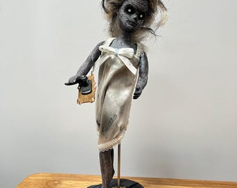 May include: A handmade doll with a spooky aesthetic. The doll features dark skin, white eyes, and grey hair. It is dressed in a white dress with a bow and holds a small, aged tag. The doll is mounted on a black base and stands on a wooden table.