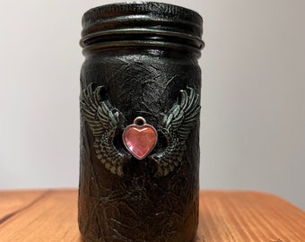 May include: A black glass jar with a textured surface and a silver charm with a pink heart hanging from it. The jar has wings on either side of the charm.