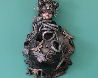 May include: A black and silver sculpture of a woman with a snake wrapped around her head and body. The woman is wearing a crown of roses and has a skull mask over her eyes. The sculpture is set against a teal background.