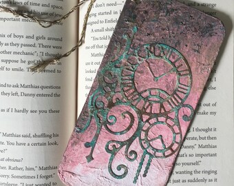 May include: A pink and green bookmark featuring a clock face design. The bookmark is made of paper and has a distressed look. The text "Danny" is written at the bottom of the bookmark.