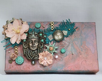 May include: A mixed media canvas artwork featuring a bronze-coloured woman's face, teal and gold embellishments, and a pink and brown background.
