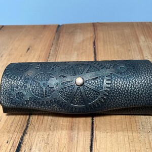 May include: A dark grey, steampunk-style glasses case with a textured surface. The case features a gear design and a small, round, copper-coloured button. The case is closed and resting on a wooden surface.