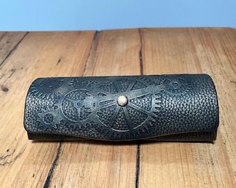 May include: A dark grey, steampunk-style glasses case with a textured surface. The case features a gear design and a small, round, copper-coloured button. The case is closed and resting on a wooden surface.