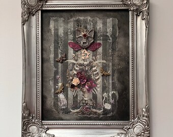 May include: A framed artwork with a silver ornate frame. The artwork features a skeletal figure with a cat-like skull, adorned with flowers and butterflies. The background has vertical stripes in shades of grey.