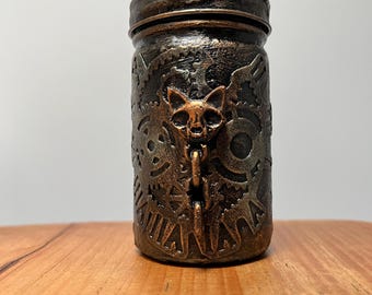 May include: A decorative jar with a steampunk design. The jar is painted in shades of bronze and copper, featuring gears and a cat skull. The jar is sitting on a wooden surface.
