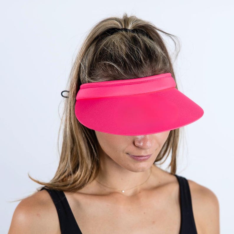 May include: A bright pink visor with a black adjustable strap. The visor is made of a soft, flexible material and has a wide brim.