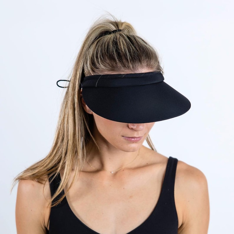 May include: A black visor with a wide brim and a black elastic band. The visor is being worn by a person with long blonde hair.