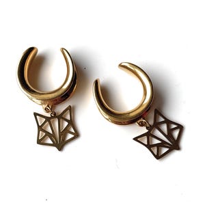 Può includere: Un paio di dilatatori per orecchie color oro con un ciondolo a forma di volpe geometrica appeso a ciascuno. I dilatatori hanno una forma curva, simile a un ferro di cavallo. I ciondoli a forma di volpe sono costituiti da sottili linee che formano una testa di volpe stilizzata.