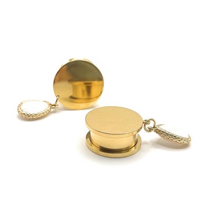 Può includere: Un paio di dilatatori per orecchie dorati con una parte superiore rotonda e piatta e una pietra rotonda bianca più piccola che pende dalla parte inferiore.