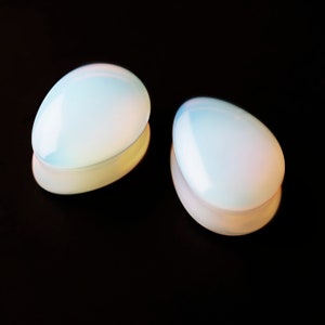 Könnte beinhalten: Zwei irisierende weiße Opal-Stöpsel, jeweils mit einer glatten, ovalen Form, werden auf einem schwarzen Hintergrund gezeigt.