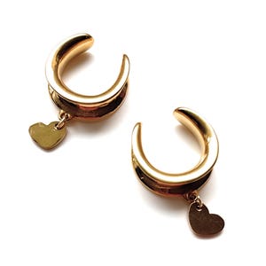 Può includere: Un paio di dilatatori per orecchie color oro con un ciondolo a forma di cuore. I dilatatori hanno una forma curva, a ferro di cavallo. Il ciondolo a forma di cuore pende da un piccolo anello nella parte inferiore del dilatatore.