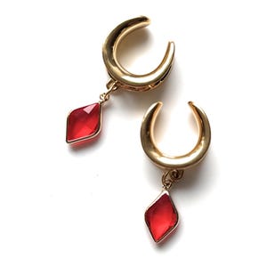 Può includere: Un paio di dilatatori per orecchie color oro a forma di mezzaluna. Ogni dilatatore ha un ciondolo rosso a forma di diamante. I dilatatori sono su uno sfondo bianco.