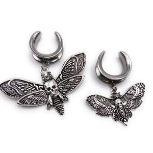 Könnte beinhalten: Ein Paar silberne Ohrdehner aus Metall mit einem baumelnden Totenkopf- und Mottiv-Design. Die Dehnungen sind aus Edelstahl und in verschiedenen Größen erhältlich.