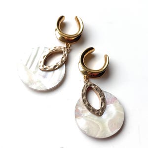 Può includere: Un paio di dilatatori per orecchie color oro con un design pendente. Ogni dilatatore presenta un ciondolo ovale martellato color oro e un ciondolo rotondo madreperlato. I dilatatori sono esposti su sfondo bianco.