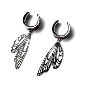 Può includere: Un paio di dilatatori per orecchie argentati con un design a forma di ala. I dilatatori hanno una forma curva, a ferro di cavallo, con un delicato ciondolo ad ala traforata che pende sotto. Le ali presentano intricati dettagli venati.