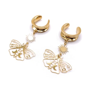 Könnte beinhalten: Ein Paar goldfarbene Ohrmanschetten mit baumelnden Anhängern. Jeder Anhänger zeigt ein zartes Schmetterlingsdesign mit Mond- und Sonnenakzenten.