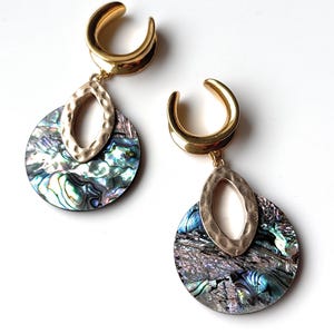Può includere: Un paio di dilatatori per orecchie color oro con un design a forma di goccia di conchiglia di abalone iridescente. Ogni dilatatore presenta un ciondolo ovale color oro martellato sospeso sotto. I dilatatori sono progettati per piercing all'orecchio allungati.