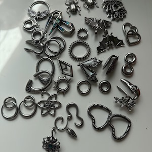 Puede incluir: Una colección de joyas corporales plateadas, que incluyen pendientes, tapones y otros adornos. El surtido presenta varios diseños como insectos, murciélagos, formas florales y patrones geométricos. Las joyas se exhiben sobre una superficie blanca.
