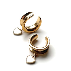 Può includere: Due dilatatori per orecchie color oro con un ciondolo a forma di cuore. I dilatatori hanno un design curvo a forma di tunnel. Ogni ciondolo a forma di cuore è bianco con un bordo dorato. Su sfondo bianco.