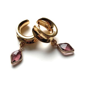 Può includere: Un paio di orecchini color oro con una gemma rosa scuro a forma di diamante pendente. Gli orecchini sono su sfondo bianco. Gli orecchini hanno un design curvo e circolare.