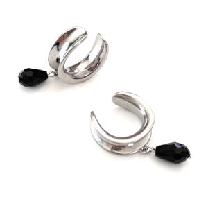 Può includere: Due orecchini a cerchio argentati con una gemma nera a forma di goccia pendente da ciascuno. Gli orecchini hanno un design curvo e sono su sfondo bianco.