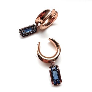 Può includere: Due orecchini a cerchio in oro rosa con pendenti rettangolari di gemme blu. Gli orecchini a cerchio hanno un design curvo e i pendenti sono incorniciati con un metallo scuro. Gli orecchini sono su sfondo bianco.