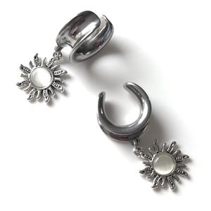 Può includere: Un paio di dilatatori per orecchie argentati con ciondoli a forma di sole. Ogni dilatatore ha un design curvo a forma di sella. Il ciondolo a forma di sole presenta un design dettagliato e radiante con una pietra centrale, circolare e bianca.