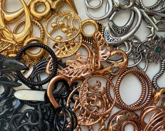 May include: A collection of decorative metal jewellery components in gold, silver, black, and copper tones. The assortment includes various shapes such as hoops, hearts, and floral designs. Some pieces feature gemstones or intricate patterns, suitable for jewellery making.
