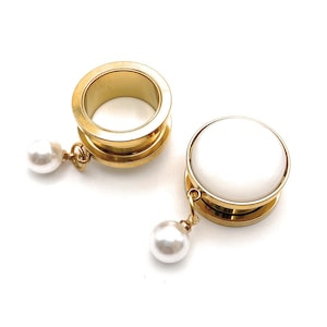 Op de afbeelding: Een paar gouden oorpluggen met een witte parel die aan elk hangt. De pluggen zijn gemaakt van roestvrij staal en hebben een gladde, gepolijste afwerking.