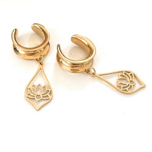 Può includere: Un paio di dilatatori per orecchie color oro. Ogni dilatatore ha un design curvo a forma di sella con un ciondolo a forma di goccia. Il ciondolo presenta un design a fiore di loto traforato. I dilatatori sono esposti su sfondo bianco.