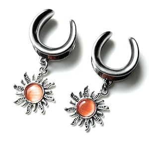 Può includere: Un paio di dilatatori per orecchie argentati con un ciondolo a forma di sole. Il ciondolo a forma di sole presenta un centro rotondo color arancione e raggi dettagliati. I dilatatori hanno un design curvo a forma di ferro di cavallo.