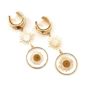 Può includere: Espansori per orecchie color oro con un ciondolo a forma di raggio di sole e un pendente rotondo trasparente contenente una margherita essiccata. La margherita ha un centro giallo e petali bianchi.