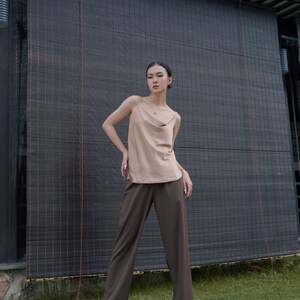 May include: A beige sleeveless camisole top with a draped cowl neckline and thin spaghetti straps paired with dark brown wide-leg trousers and white pointed-toe shoes. The outfit features a neutral colour palette and a clean, simple silhouette.