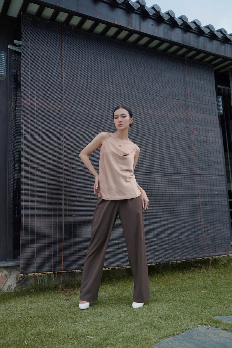 May include: A beige sleeveless camisole top with a draped cowl neckline and thin spaghetti straps paired with dark brown wide-leg trousers and white pointed-toe shoes. The outfit features a neutral colour palette and a clean, simple silhouette.