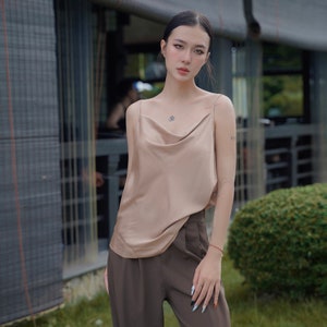 May include: A beige satin camisole featuring a draped cowl neckline and thin spaghetti straps, paired with high-waisted dark brown trousers. The camisole has a smooth, reflective finish, while the trousers feature a tailored fit with visible pleats.